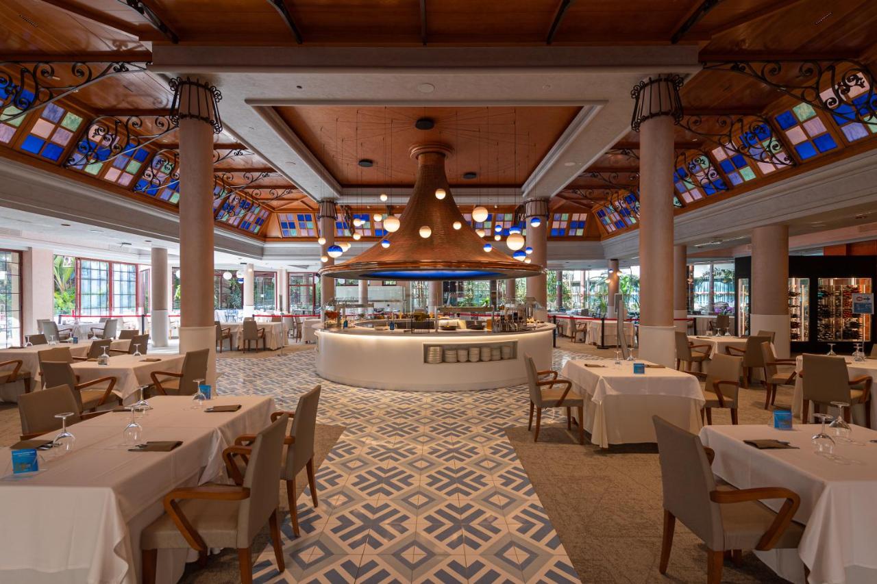 een restaurant met witte tafels en stoelen en een kroonluchter bij Hotel Cordial Mogán Playa in Puerto de Mogán