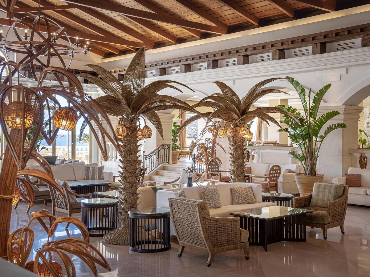 een lobby met palmbomen, stoelen en tafels bij Grecotel Marine Palace & Aqua Park in Panormos Rethymno